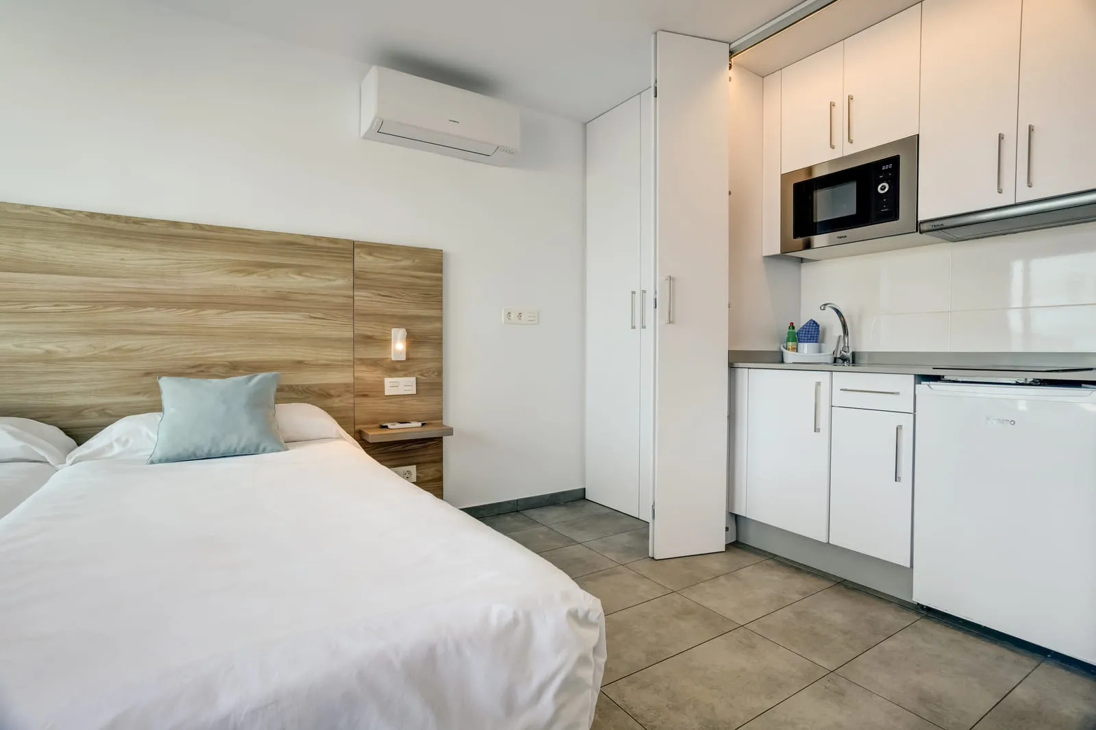 La cocina en la pared del fondo — placa, nevera, fregadero y microondas — con la cama a unos pasos